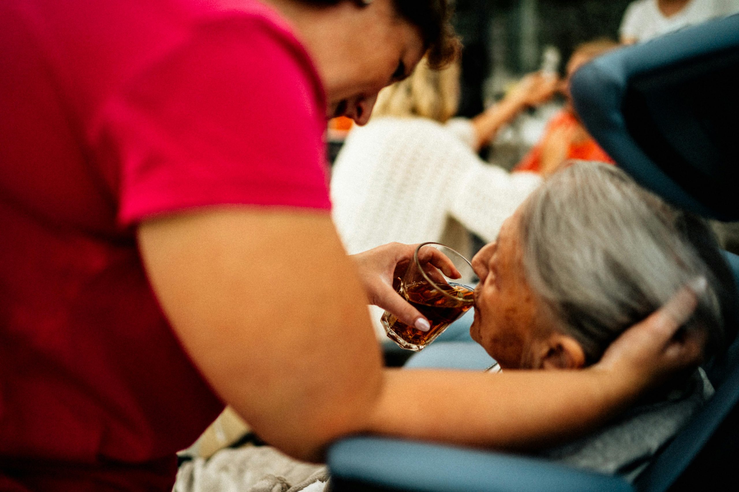 Hol di toi gUG | Soziale Hilfe, Integration & Gemeinschaft A caregiver helps an elderly woman drink tea, showcasing empathy and support.