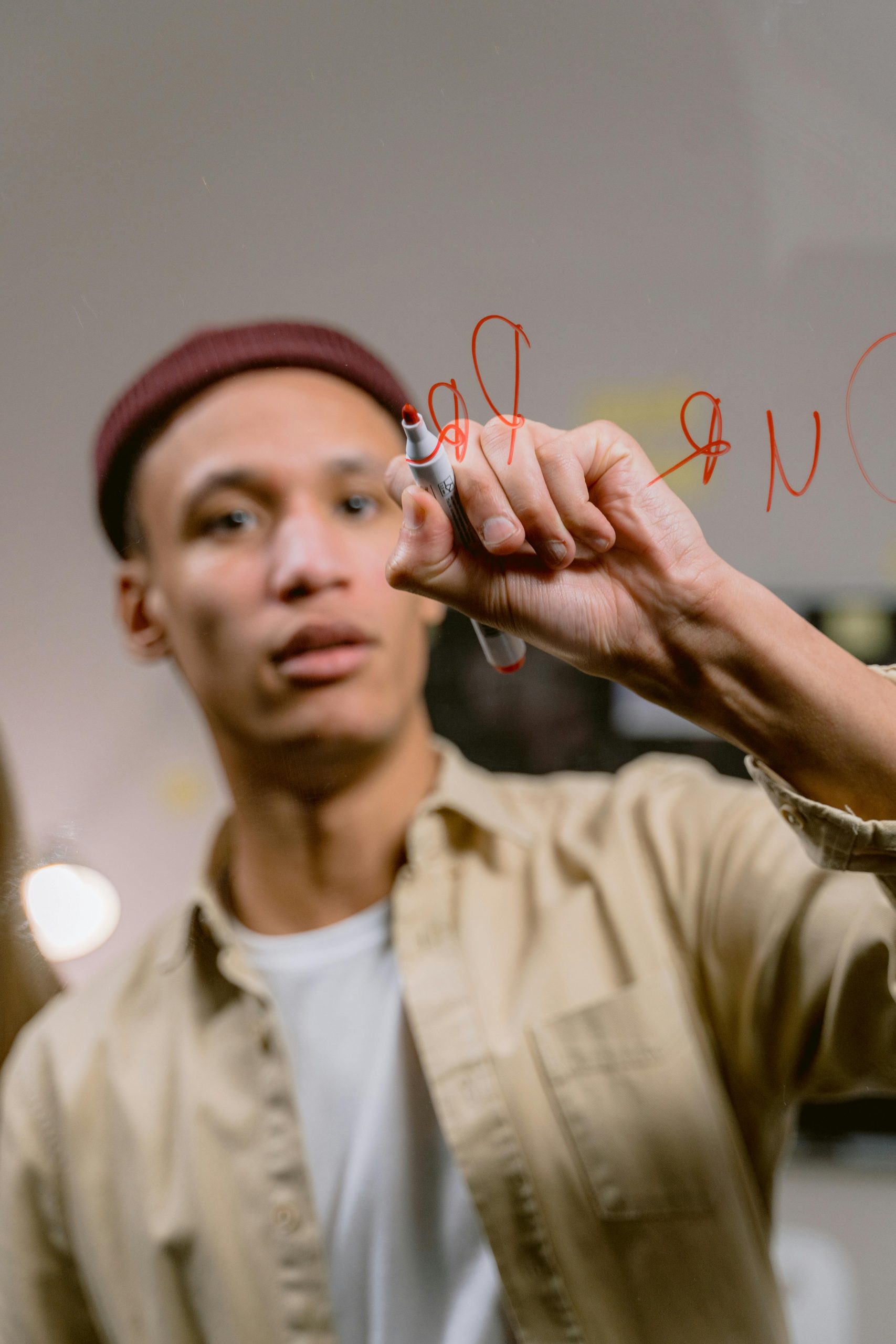 Hol di toi gUG | Soziale Hilfe, Integration & Gemeinschaft Casual dressed man wearing a beige shirt and beanie writing on clear glass with a red marker.